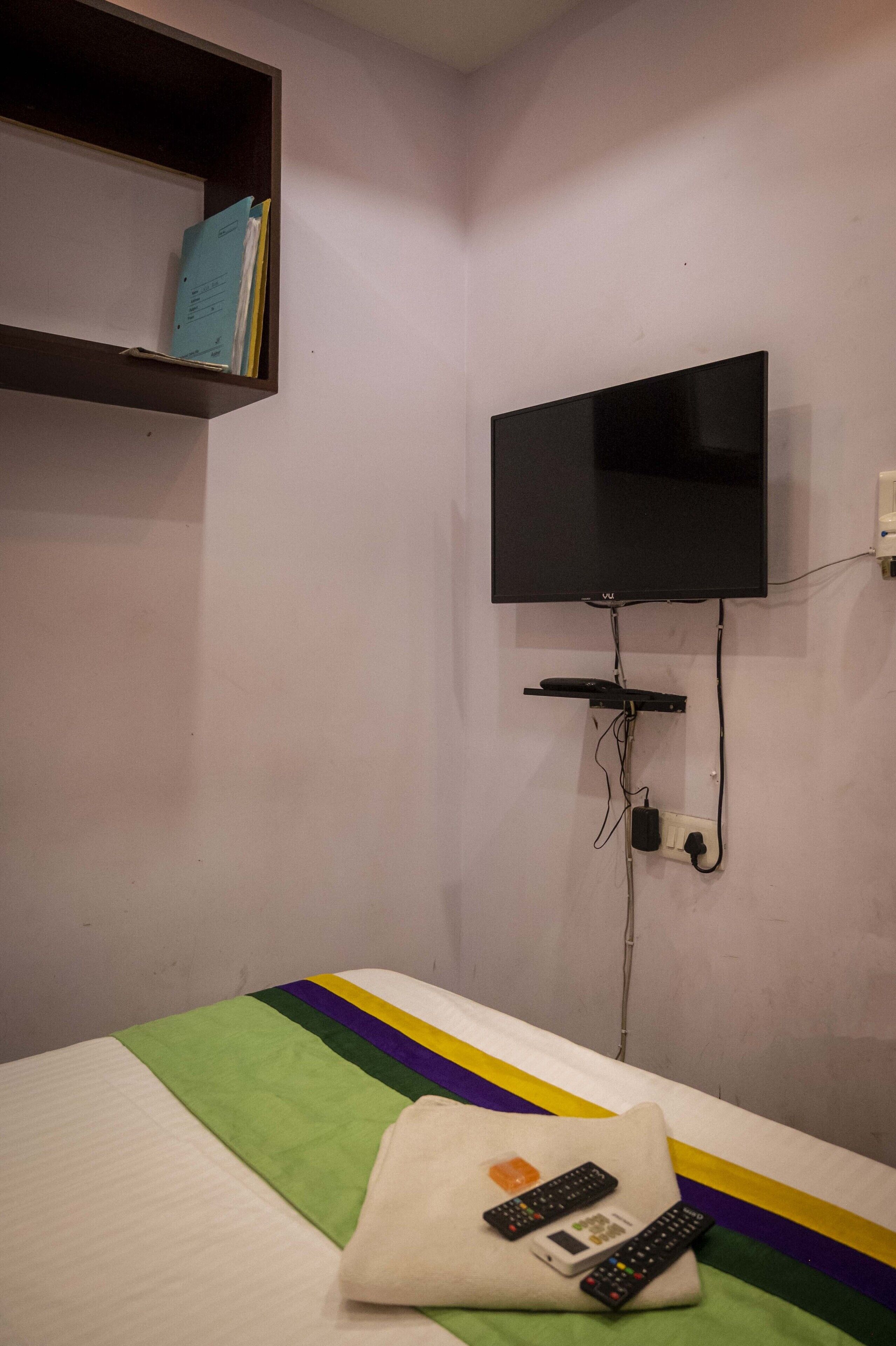 Un lit simple recouvert d’une couverture à rayures vertes et blanches, avec un oreiller et deux télécommandes posées dessus. Un téléviseur à écran plat est fixé au mur, au-dessus du lit.