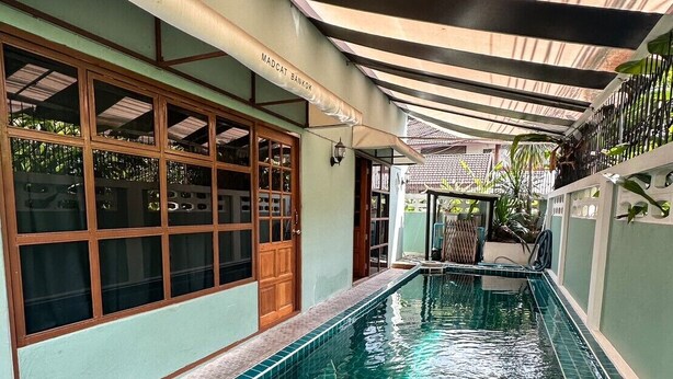 Una piscina con bordes de azulejos, una zona cubierta con techo, y un edificio con amplias ventanas y una puerta.