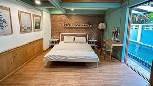 Un dormitorio con una cama grande, una pared de ladrillo, piso de madera, una pequeña mesa y vista a una piscina.