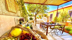 Un cesto di frutta e verdura fresca su un patio con tavolo e sedie, circondato da piante in vaso e un edificio con pareti gialle.
