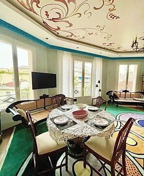 A dining area with a round table set for four, surrounded by chairs. There is a flat-screen TV mounted on the wall, and the room has large windows with curtains.