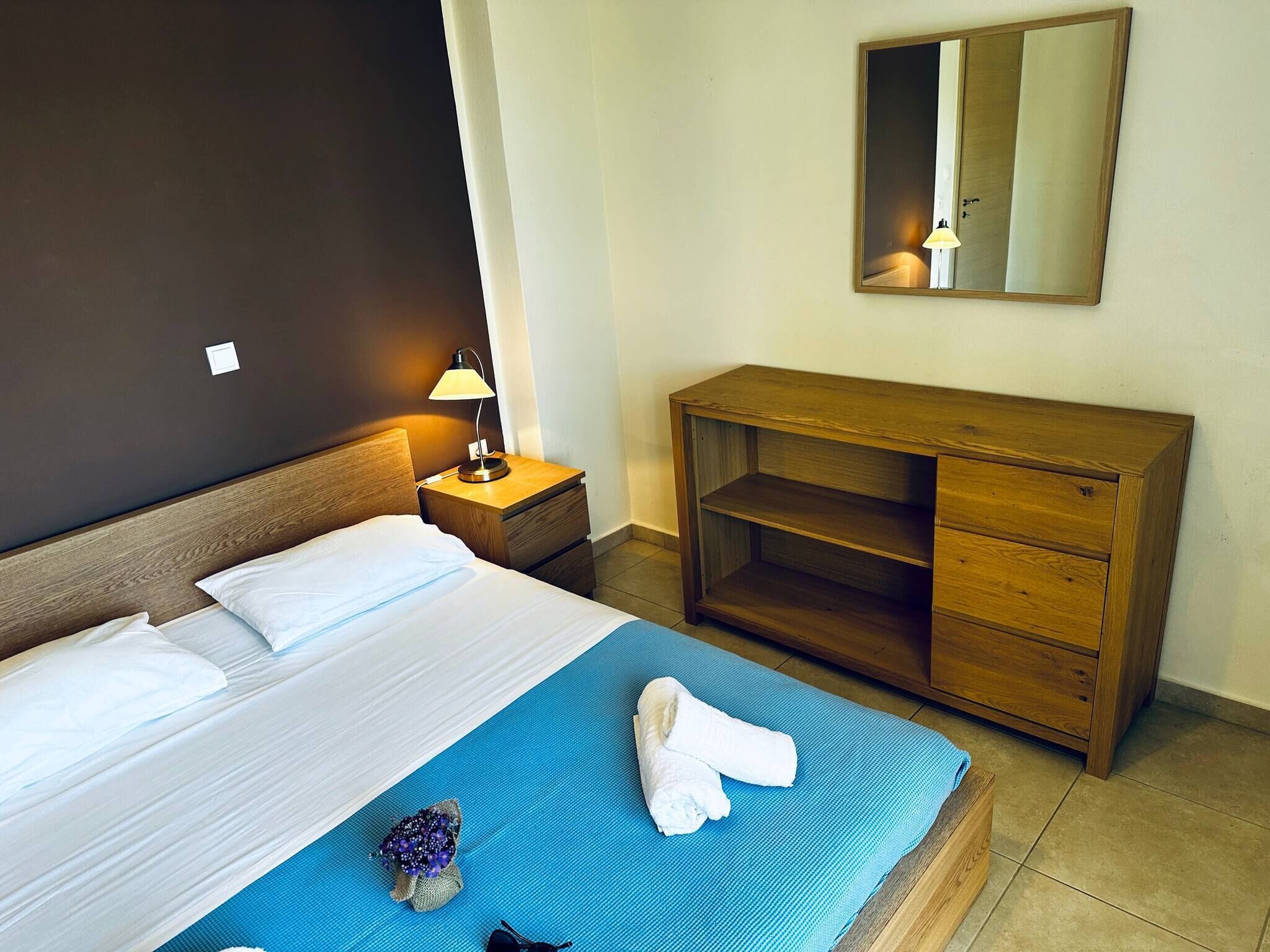 Una camera d'albergo con un letto, un comodino con una lampada, una cassettiera in legno e uno specchio sulla parete.