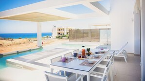 A modern outdoor dining area with a pool, white furniture, and a view of the ocean. - Starlight Villas-8bd, Available For Events (Rethymno)