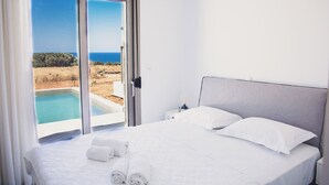 A hotel room with a bed, white linens, a nightstand, a lamp, and a view of a pool and landscape. - Starlight Villas-8bd, Available For Events (Rethymno)