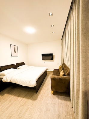 A hotel room with a bed, a sofa, a TV mounted on the wall, and a window with curtains.