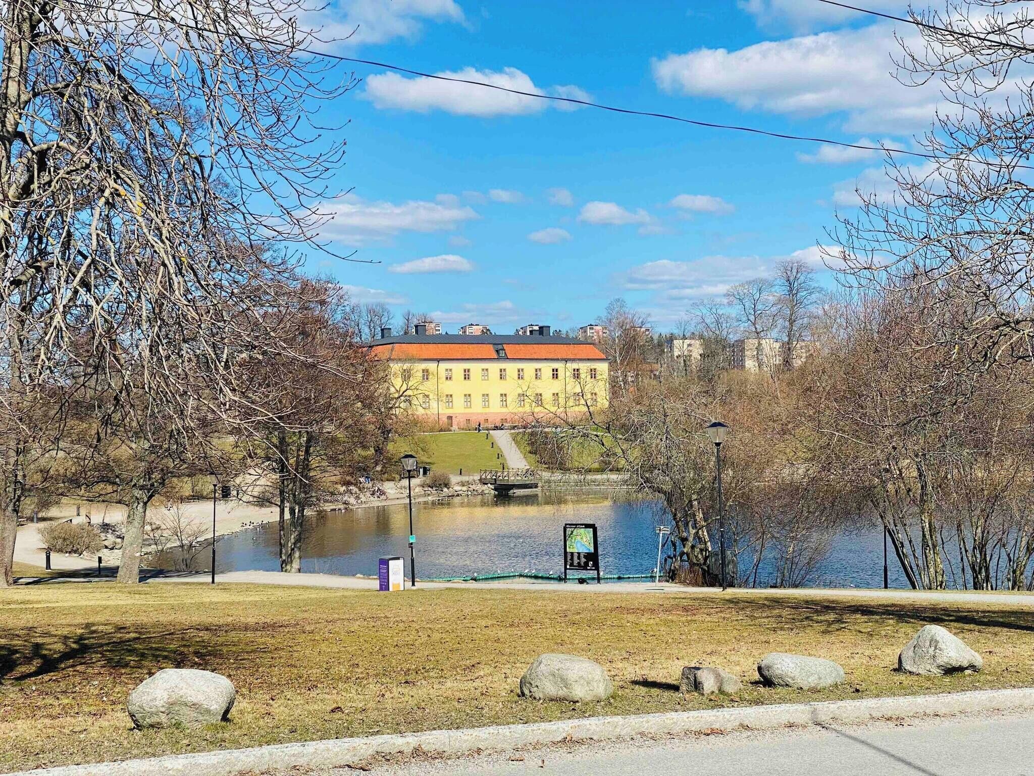 A park with a lake, a yellow building, and a bridge.