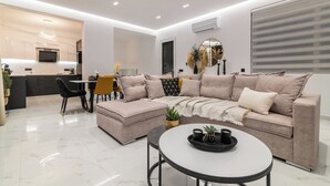 A modern living room with a sofa, ottoman, and coffee table, a dining area with chairs and a table, and a kitchen with a TV and counter. - Melissia Luxury Villa (Zakynthos)