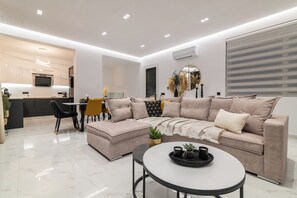 A modern living room with a sofa, ottoman, and coffee table, a dining area with chairs and a table, and a kitchen with a TV and counter.