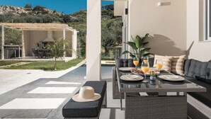 A modern outdoor dining area with a table set for breakfast, a sofa, and a pergola with a grill. - Melissia Luxury Villa (Zakynthos)