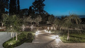 A pathway leading to a building with a view of the ocean, surrounded by trees and illuminated by lights. - Melissia Luxury Villa (Zakynthos)
