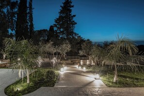 A pathway leading to a building with a view of the ocean, surrounded by trees and illuminated by lights.