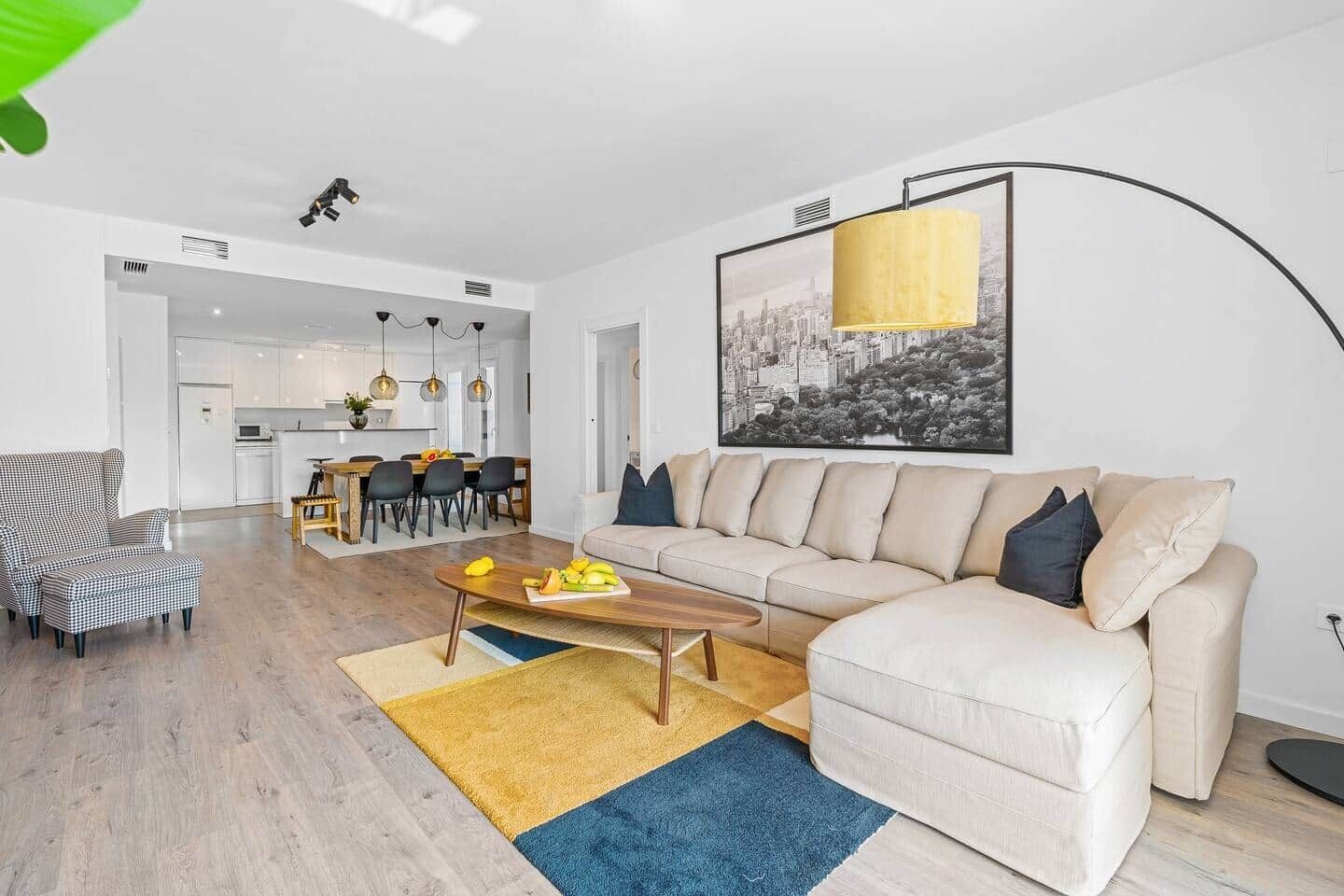 A modern living room with a beige sofa, a checkered armchair, and a coffee table. There is a dining area in the background with a kitchen visible.