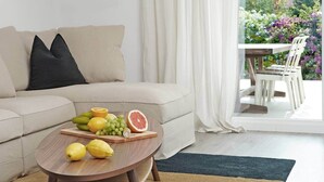 Un salon avec un canapé beige, une table basse en bois sur laquelle sont disposés des fruits, et une vue sur un jardin par la fenêtre.