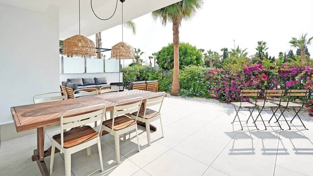Un espace repas extérieur moderne, avec une table et des chaises en bois, entouré d’une végétation luxuriante et de fleurs.