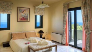A bedroom with a bed, a bedside table, a lamp, a painting, and a view of the sea through a window. - Koralia - Chania's Card Postal Villa Majestic View (Chania)