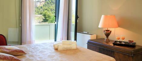 Quarto de hotel com cama, mesa de cabeceira e luminária, e vista para o verde lá fora.