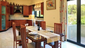 A dining area with a table set for four, wooden cabinets, a window with curtains, and a door leading to an outdoor area. - Koralia - Chania's Card Postal Villa Majestic View (Chania)