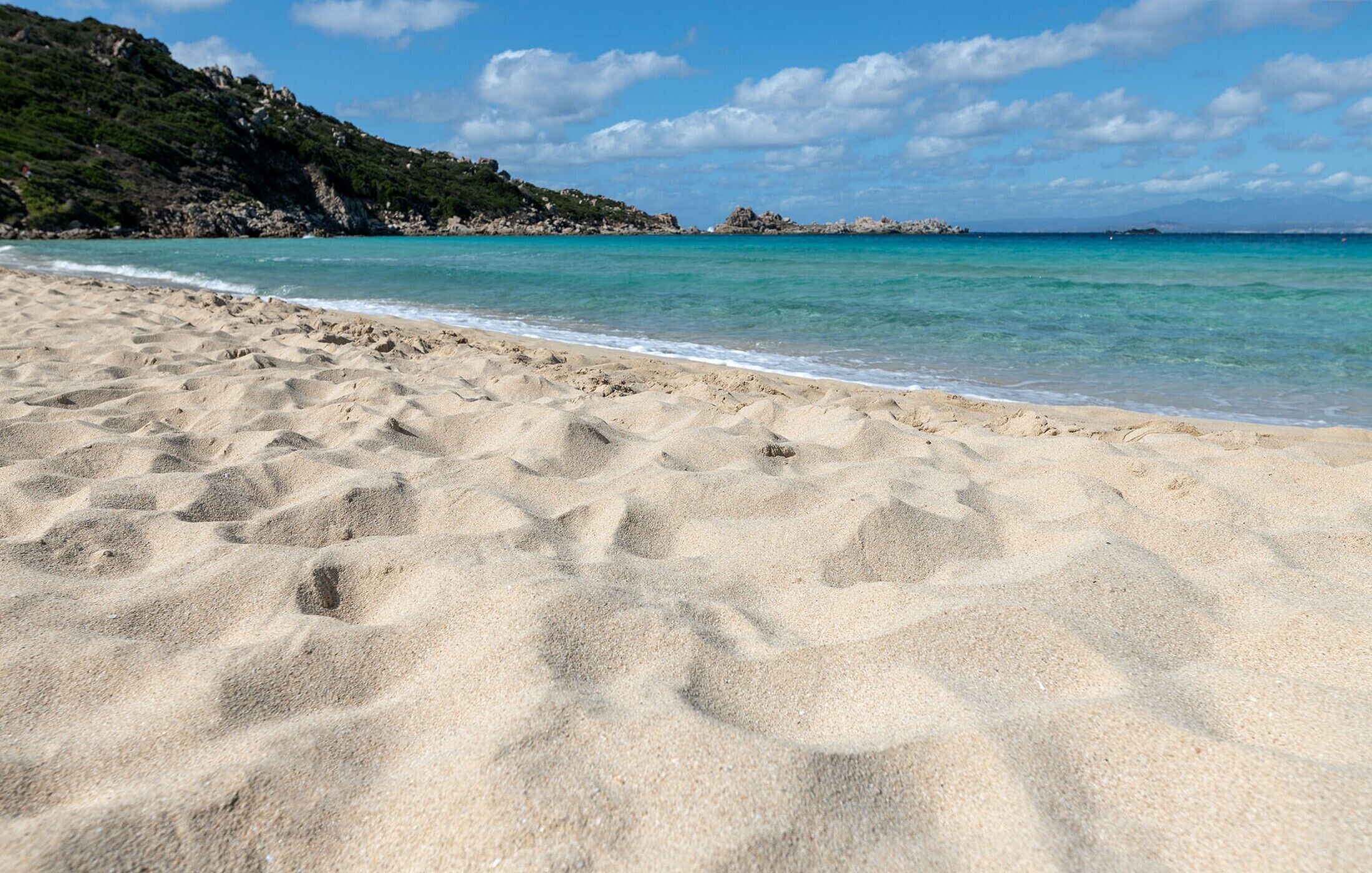Une plage de sable fin bordée d’une eau turquoise et limpide, avec des formations rocheuses et, en arrière-plan, des collines verdoyantes.
