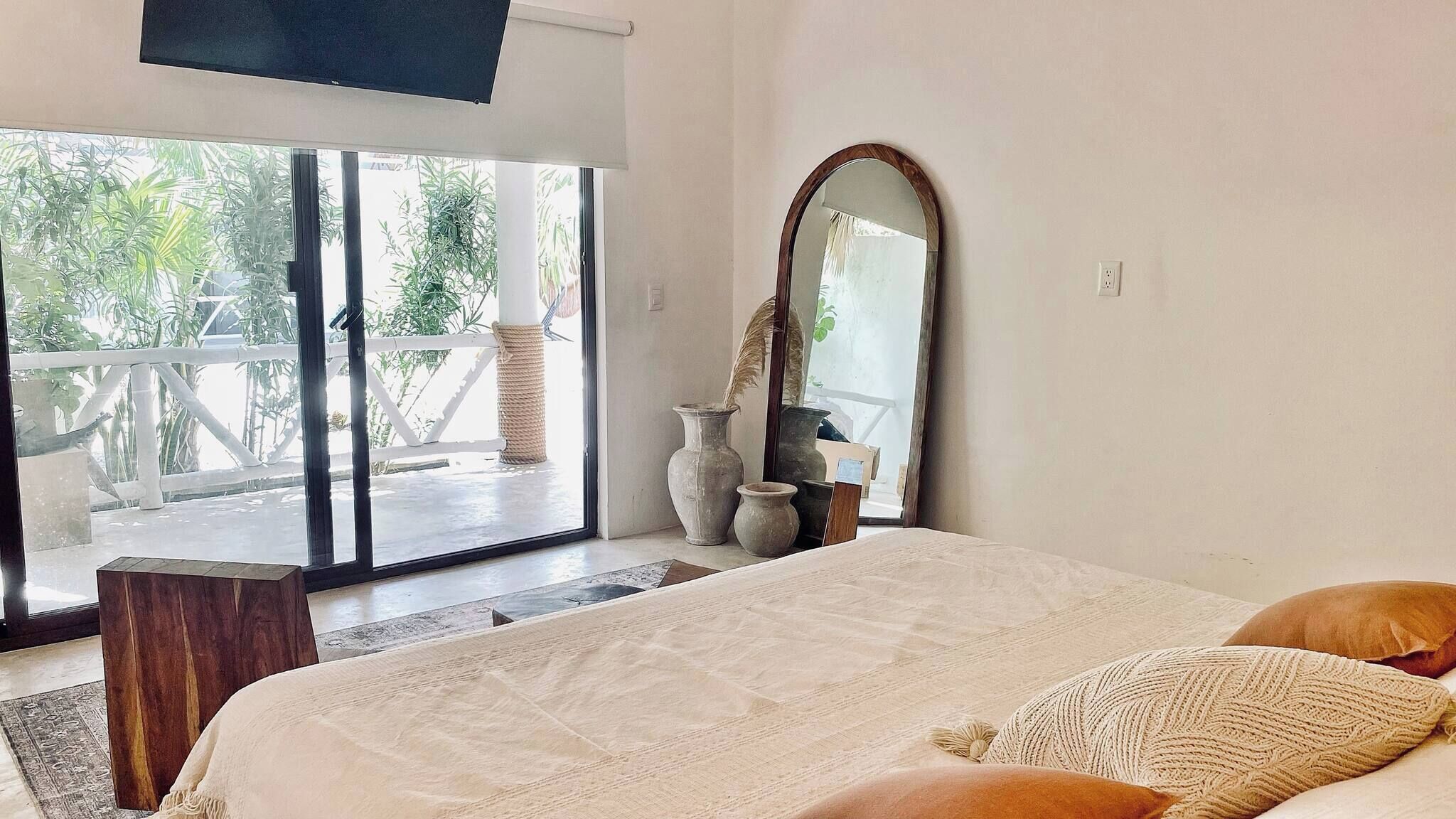 A bedroom with a large bed, a mirror, and a view of a balcony with plants.