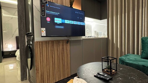 Un soggiorno moderno con una TV a schermo piatto montata a parete, un tavolino rotondo in marmo con un telecomando e fazzoletti, un divano verde e una lavatrice sullo sfondo.
