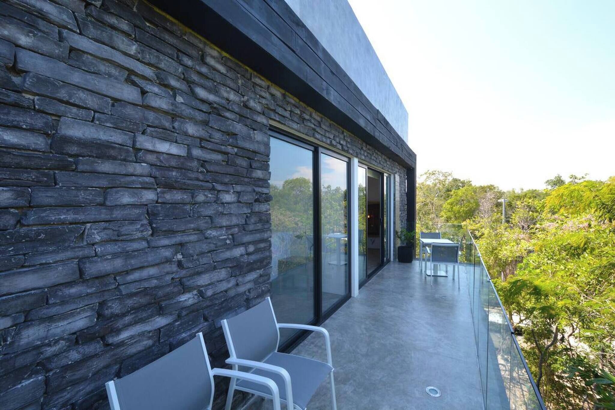 A modern building with a stone facade and large glass doors leading to a balcony with outdoor seating.