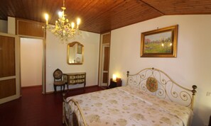 A bedroom with a wooden ceiling, a chandelier, a bed with a floral bedspread, a dresser, a chair, a mirror, and a framed landscape painting. - Spacious Villa With Garden by the sea (Lignano Sabbiadoro)