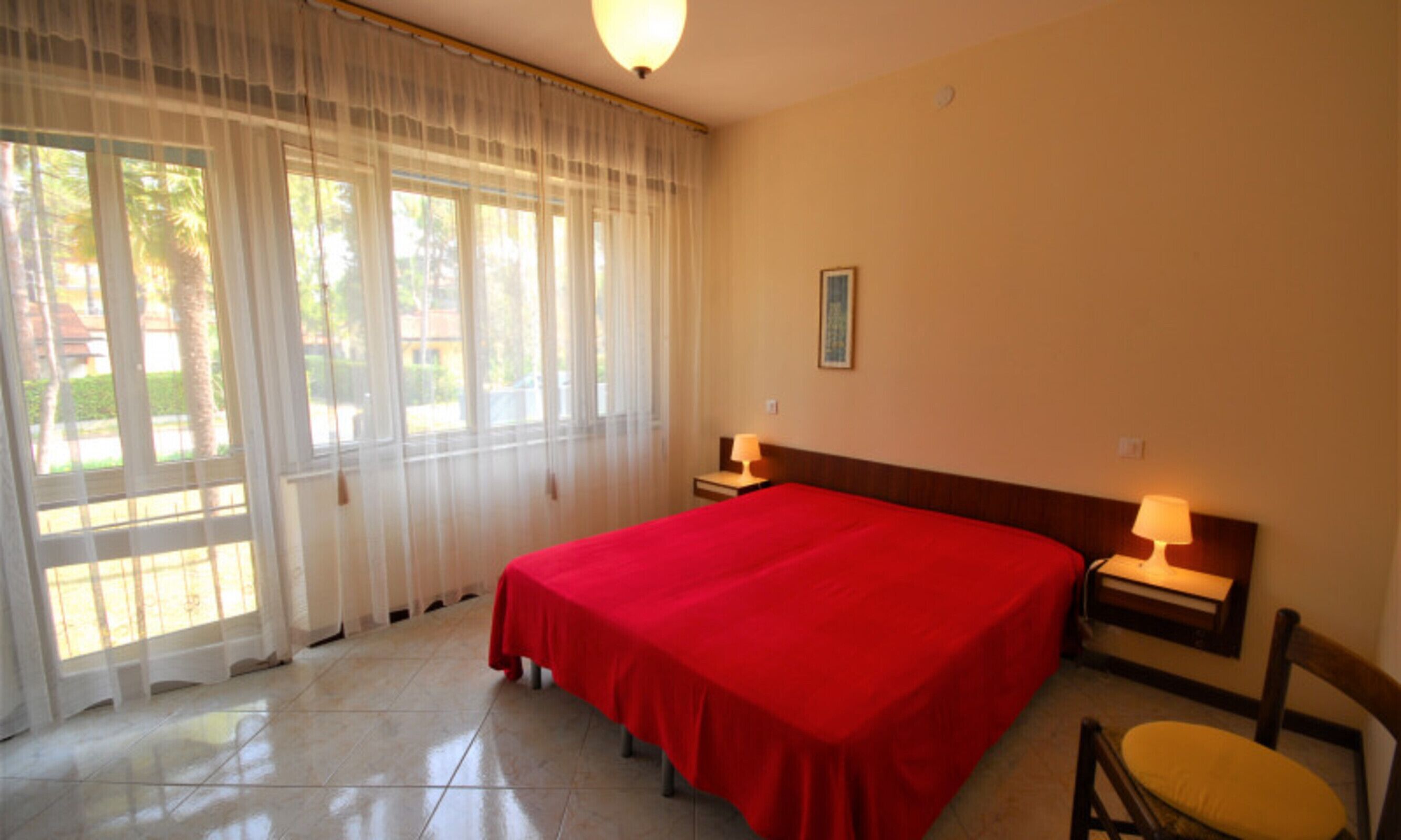 A hotel room with a large bed, two bedside tables with lamps, a chair, and a window with sheer curtains.