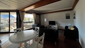 En stue med spisebord, stole, sofa, fjernsyn og en balkon med udsigt til bygninger og bjerge.