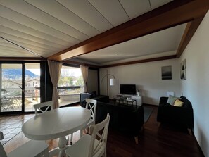 Ein Wohnzimmer mit Esstisch, Stühlen, einer Couch, einem Fernseher und einem Balkon mit Blick auf Gebäude und Berge.