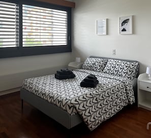 Un dormitorio con cama, mesita de noche, ventana con persianas y dos cuadros en la pared.