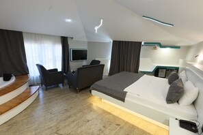 Una habitación de hotel moderna con un escritorio de madera curvada, una cama grande con ropa blanca, un sofá y un televisor de pantalla plana montado en la pared.