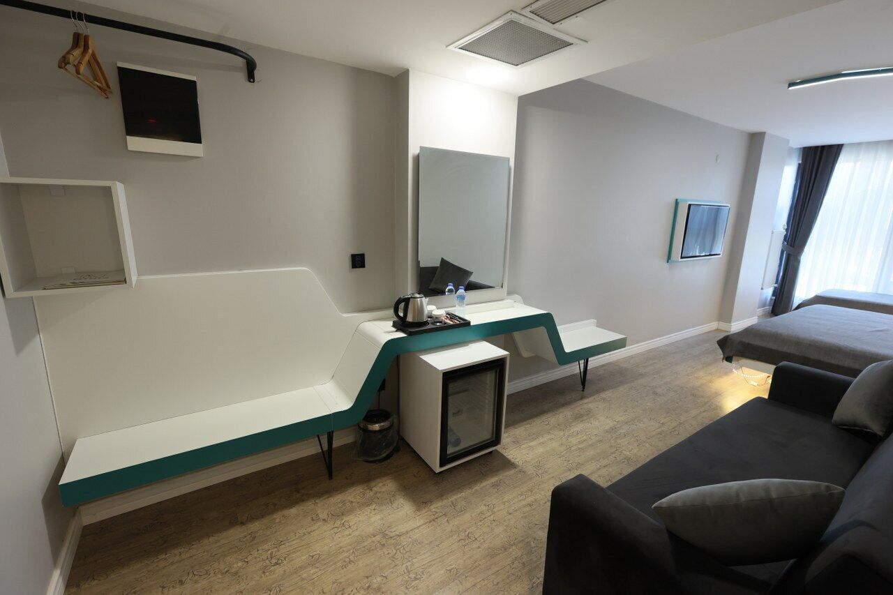 A modern hotel room with a bed, a sofa, a desk with a computer, a small TV, and a mirror.