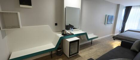 A modern hotel room with a bed, a sofa, a desk with a computer, a small TV, and a mirror.