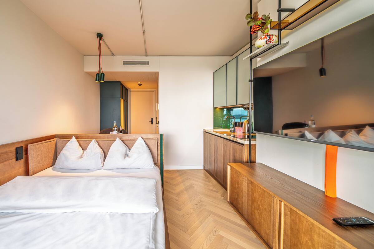 Camera d'albergo moderna con un letto, una scrivania in legno, un angolo cottura e un bagno visibile attraverso una porta aperta.