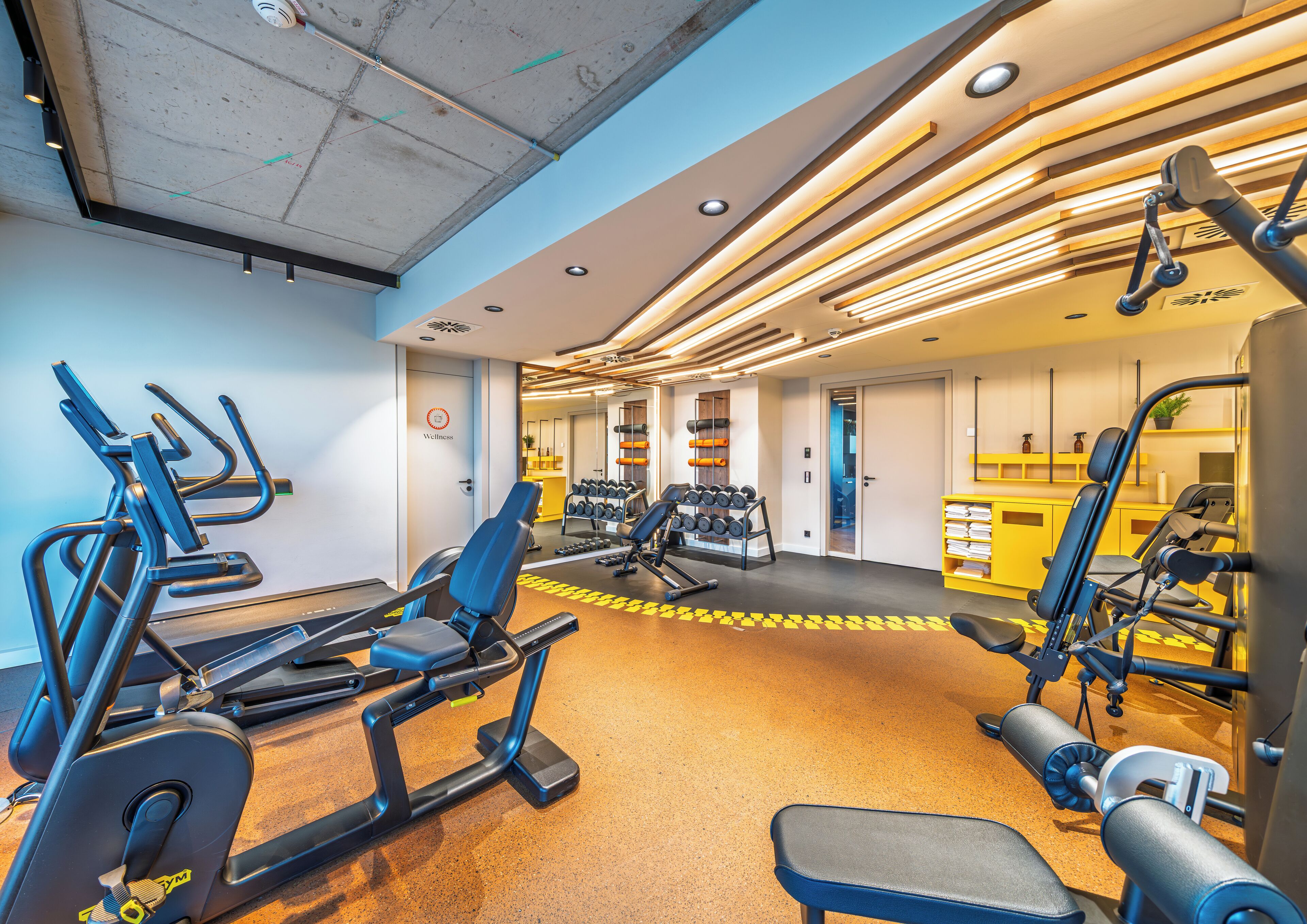 A gym with exercise equipment, a yellow wall, and a door labeled 'Wellness'.