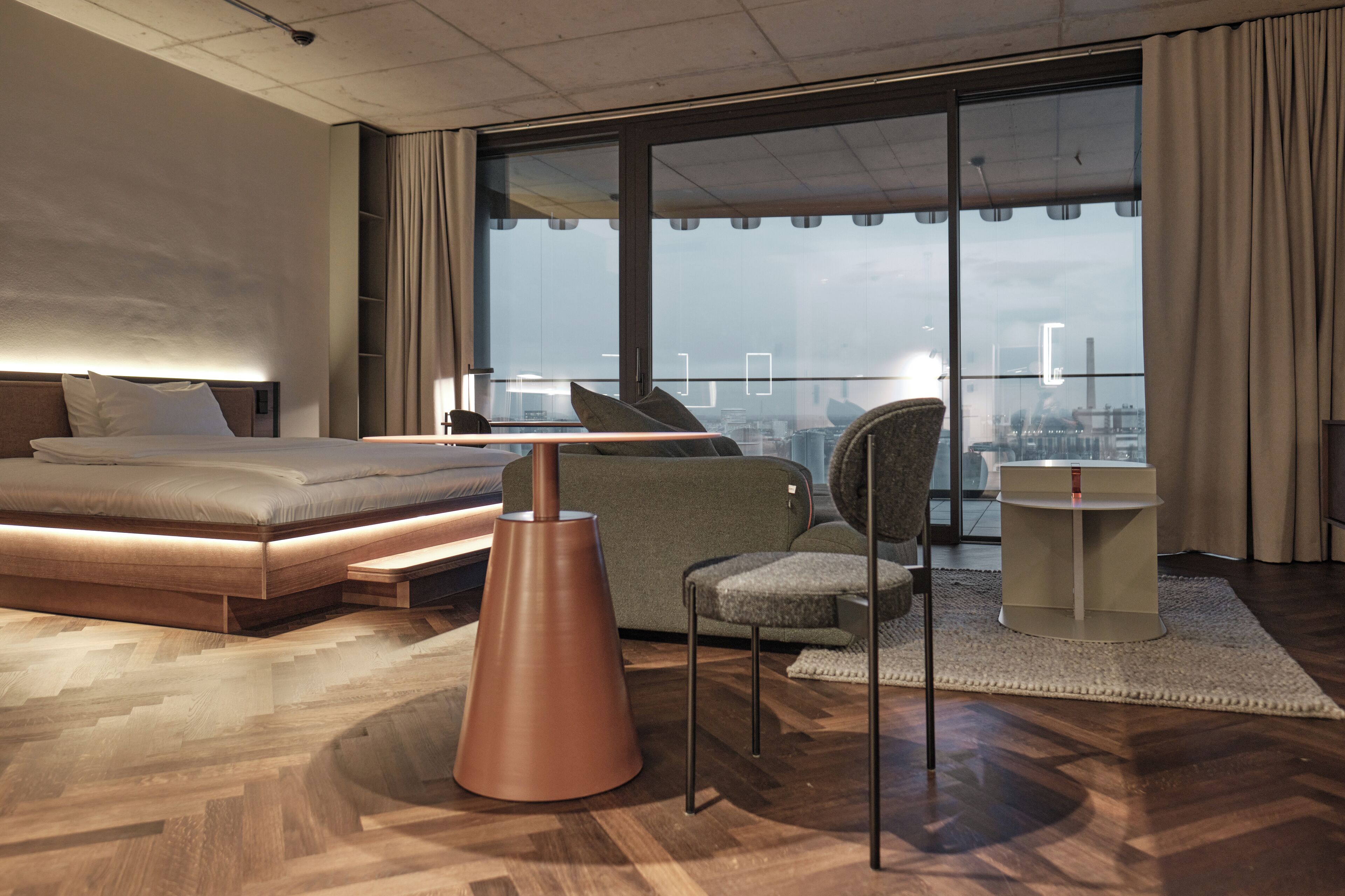 Ett modernt hotellrum med en stor säng, en soffa, ett litet bord och utsikt över staden genom stora fönster.