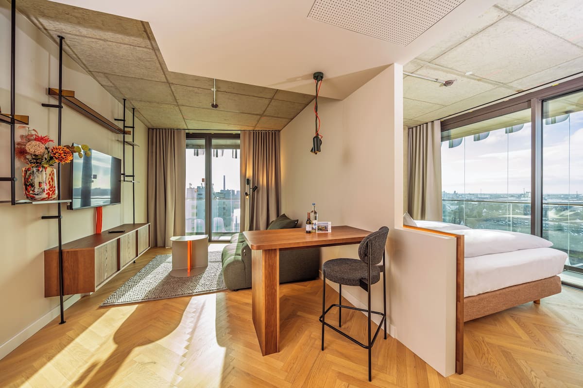 Una camera d'albergo moderna con una grande finestra, una televisione a schermo piatto, un tavolo da pranzo in legno, un divano e un letto.