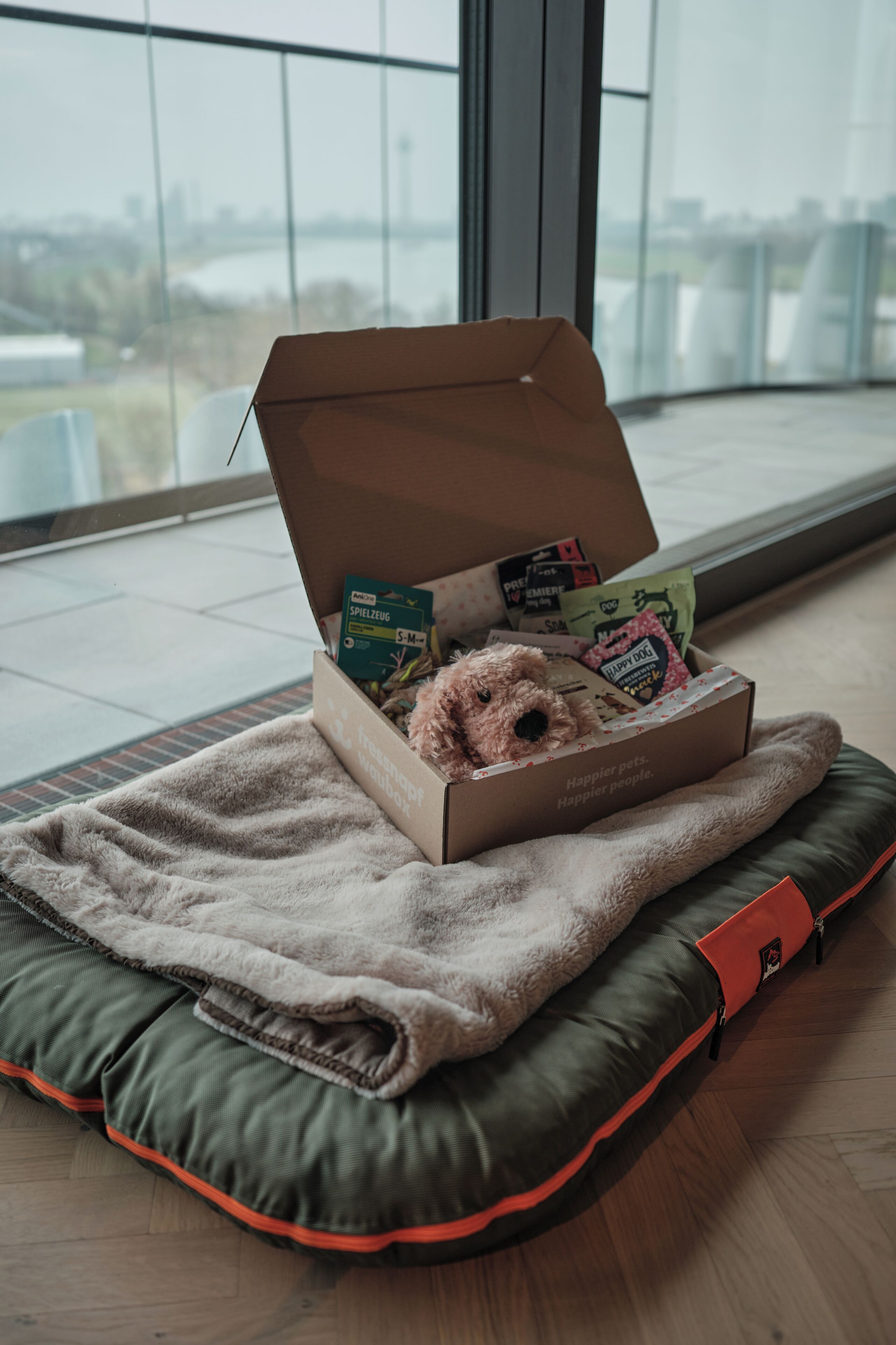 A cozy pet bed with a plush toy and pet food boxes in a room with a large window.