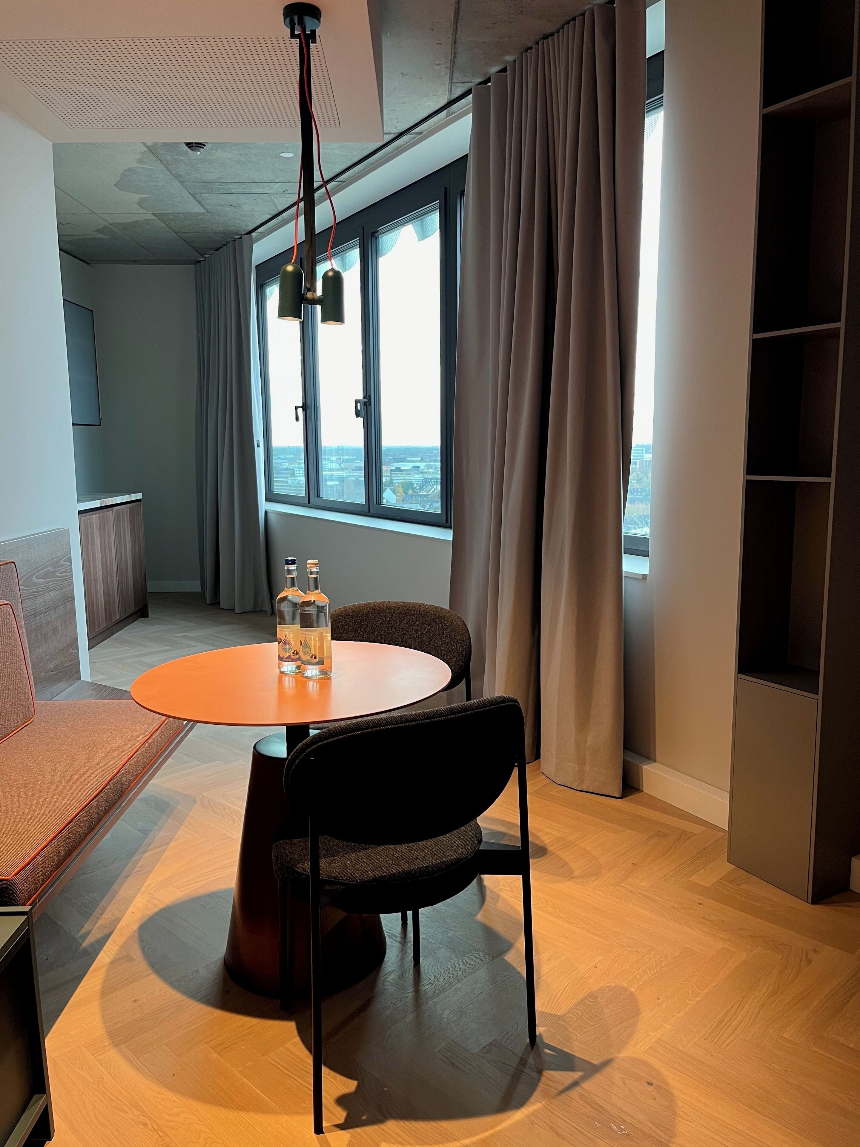 A modern room with a round table, two chairs, a sofa, a large window with curtains, and a view of the city.