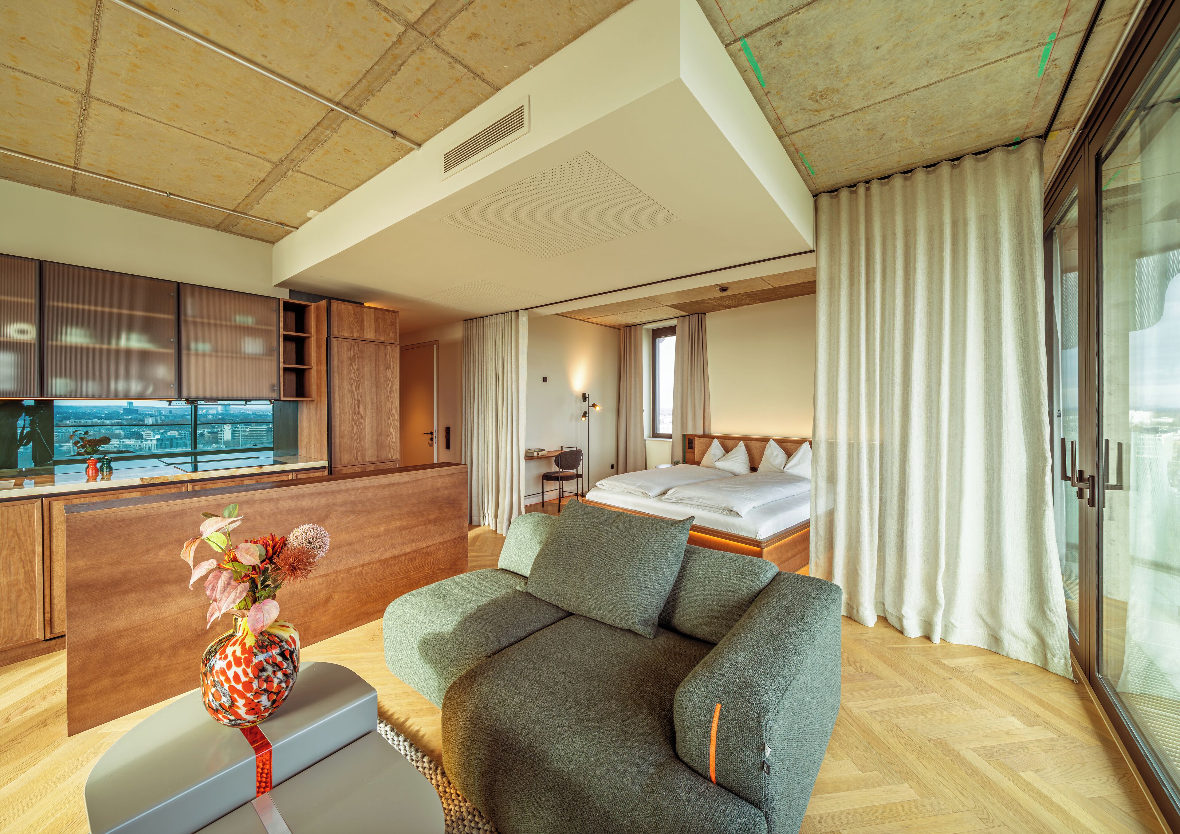Una habitación de hotel moderna con cama, sofá, una mesita con un florero, kitchenette y un amplio ventanal con cortinas.