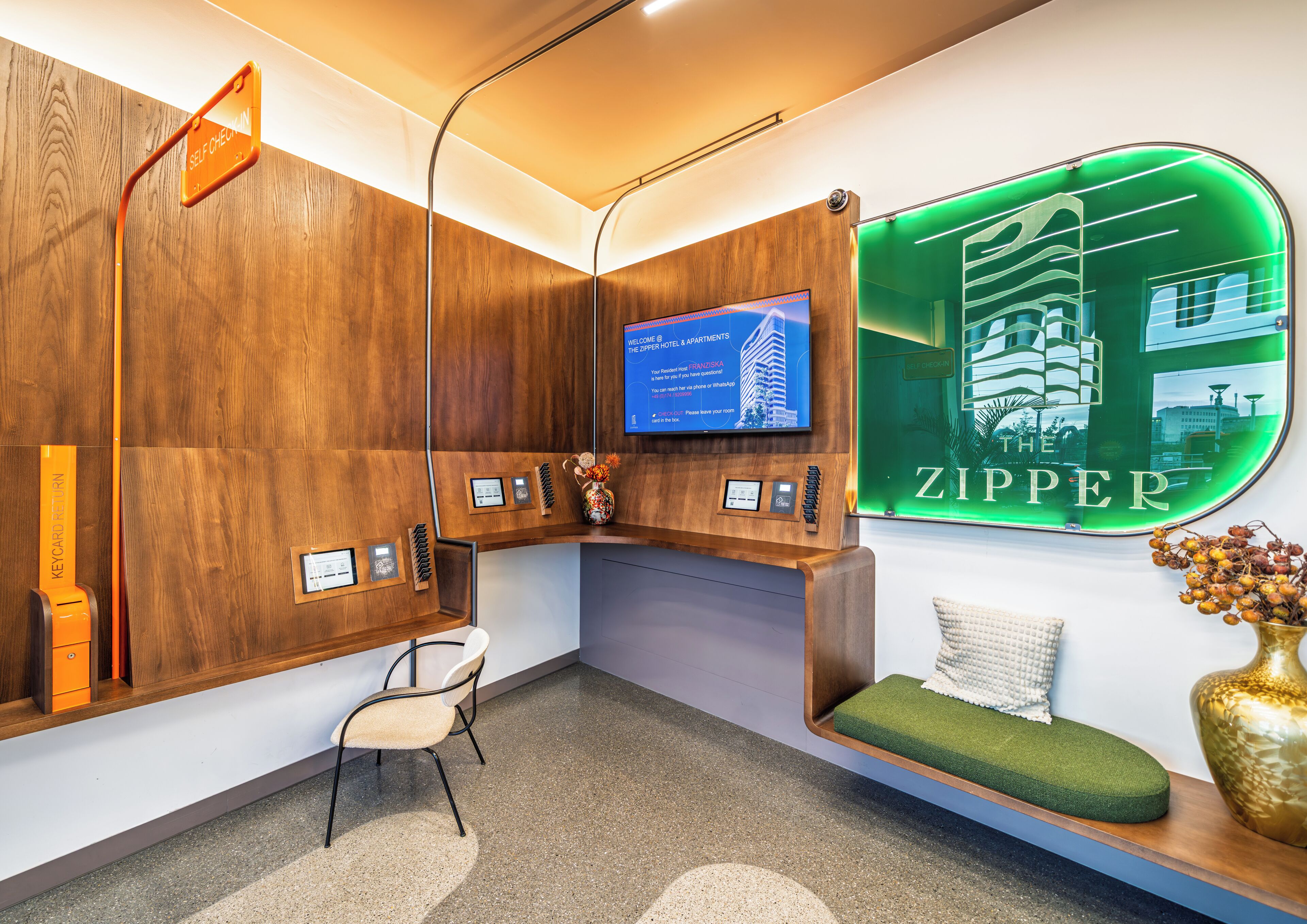 A reception area with a wooden reception desk, a television screen, a green neon sign, a seating area with a green cushion, and a wall-mounted keycard return station.