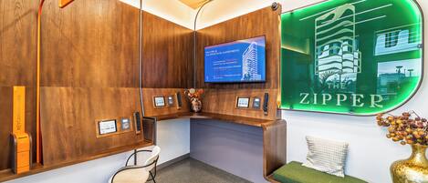 A reception area with a wooden reception desk, a television screen, a green neon sign, a seating area with a green cushion, and a wall-mounted keycard return station.
