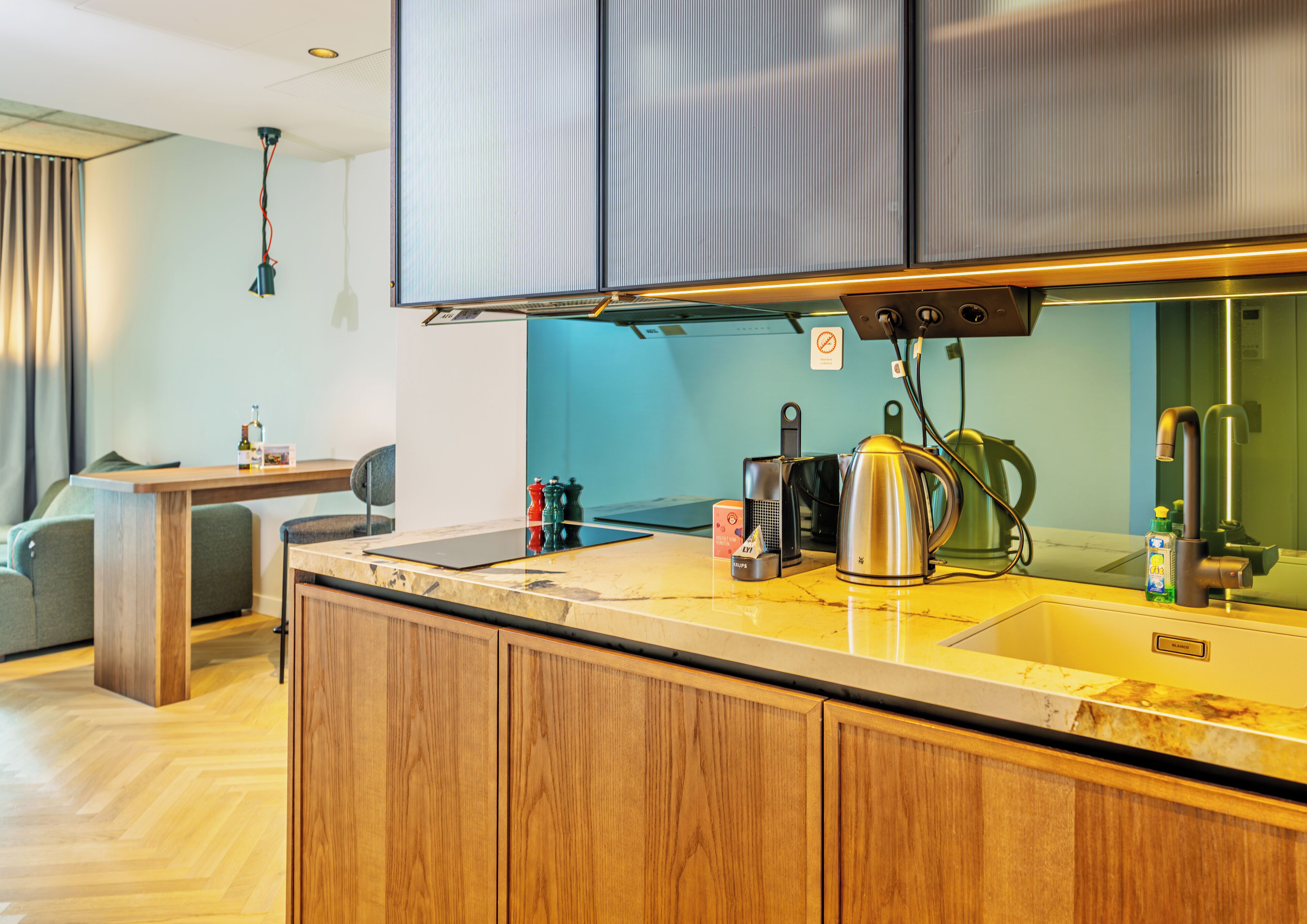 A modern kitchen with wooden cabinets, a stainless steel kettle, a coffee maker, and a small table with a chair.