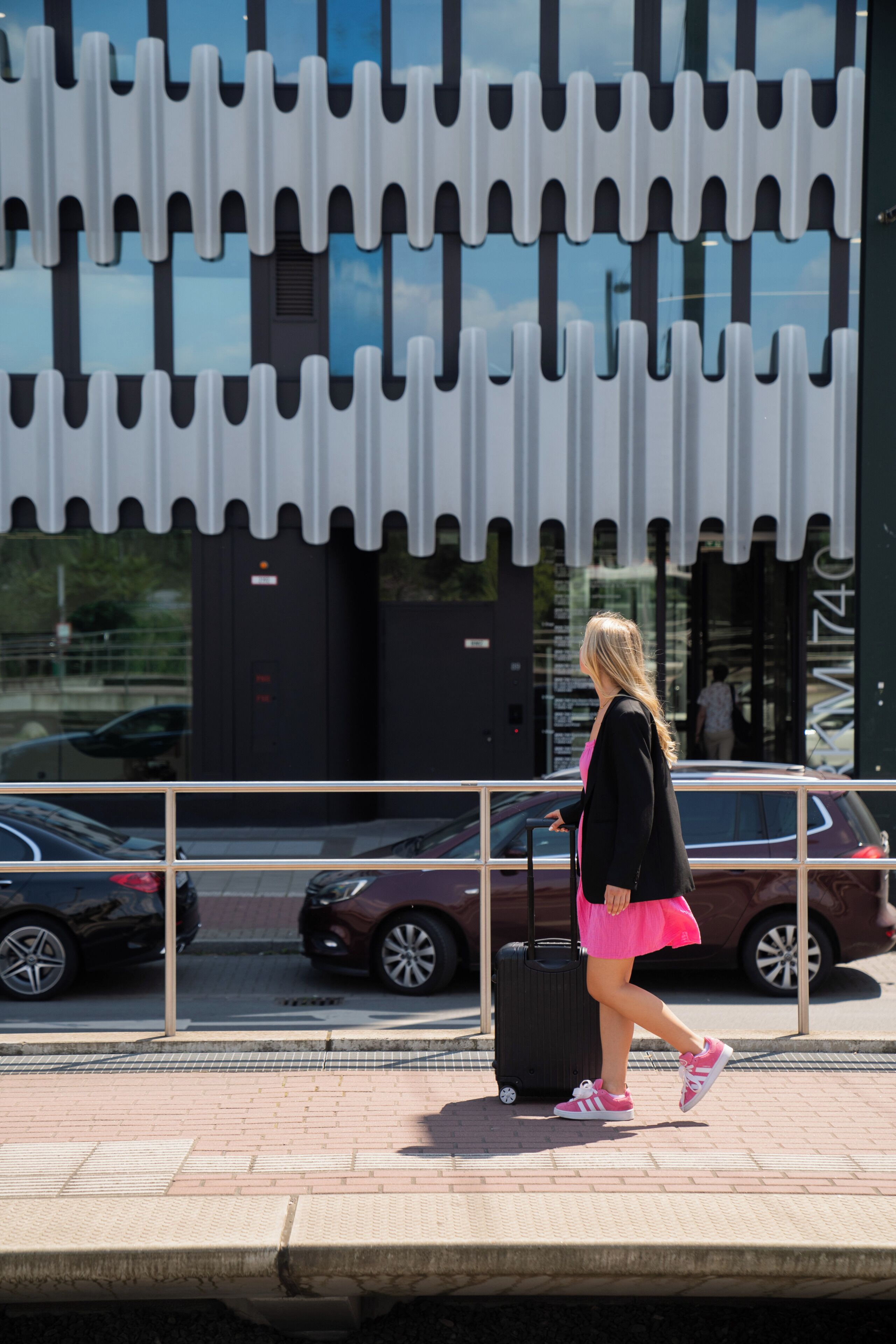 A woman walking with a suitcase on a sidewalk, passing parked cars and a building with a distinctive awning.