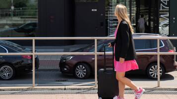 A woman walking with a suitcase on a sidewalk, passing parked cars and a building with a distinctive awning.