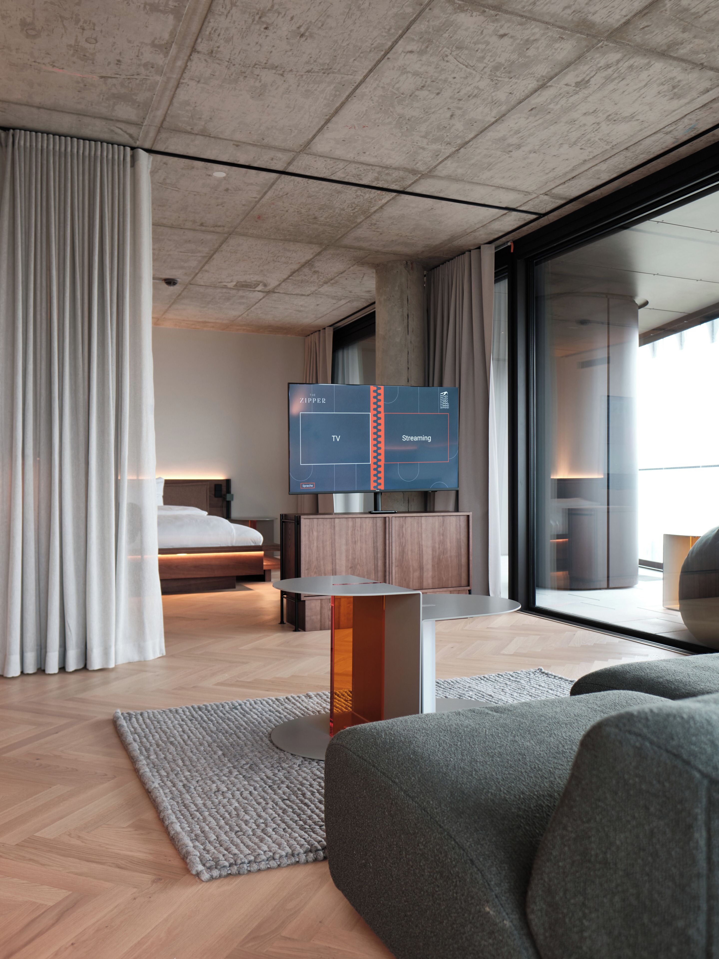 Habitación de hotel moderna con una televisión de pantalla plana grande, una cama, un sofá y vistas a la ciudad a través de amplios ventanales.