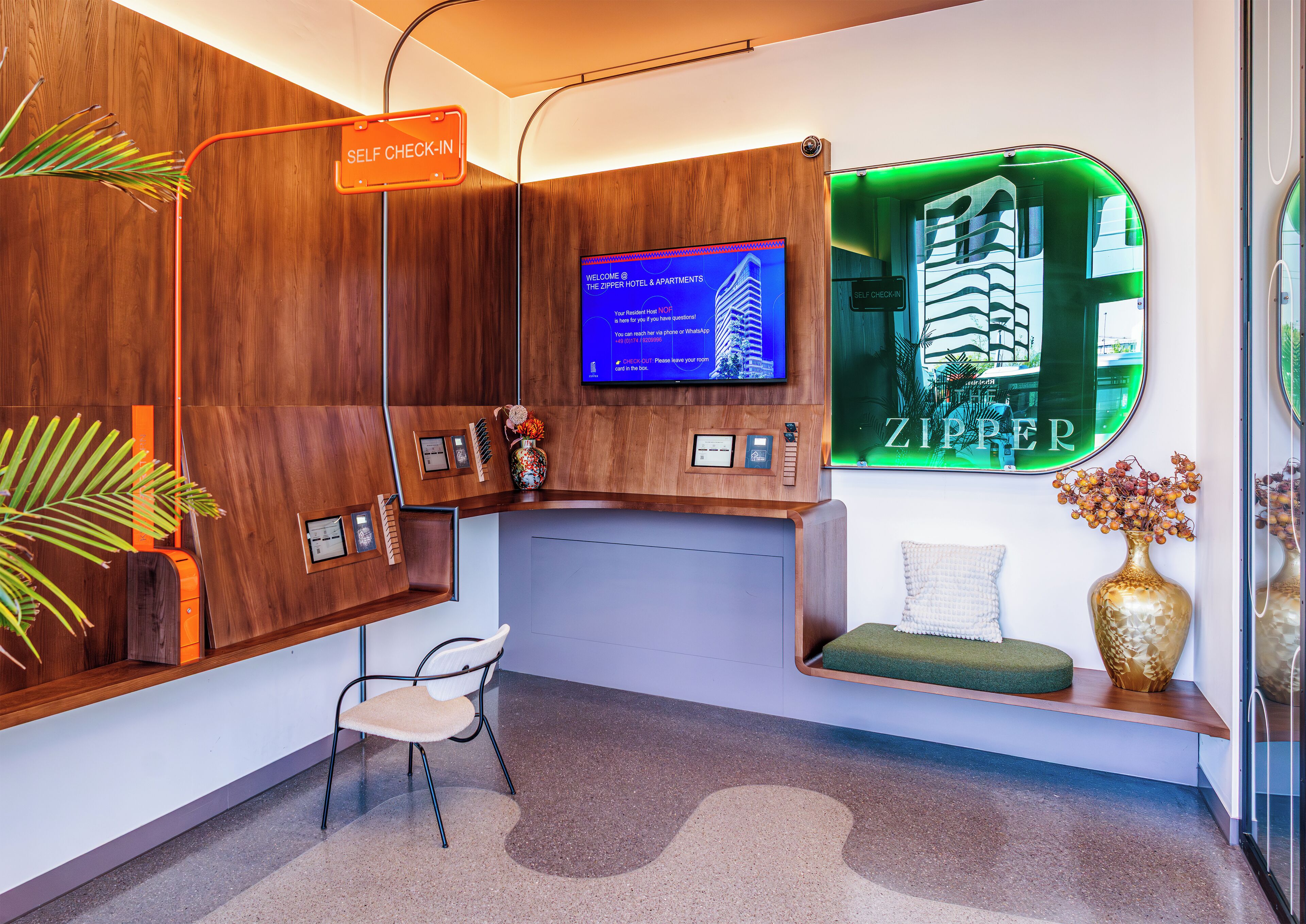 A reception desk with a self-check-in sign, a television displaying welcome messages, and a framed view of a building.