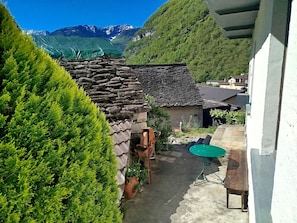 A stone building with a sloped roof, a green table, and potted plants in a courtyard with a mountain view.