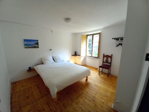 A bedroom with a wooden floor, a bed with white bedding, a window with a view of greenery, and a chair. - Room-"casa Coerente" Cavergno - Room #3 (Cevio)
