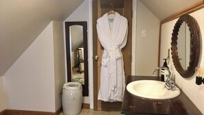 Un baño con un lavamanos blanco, tocador de madera, un espejo grande y una bata colgada en una puerta.